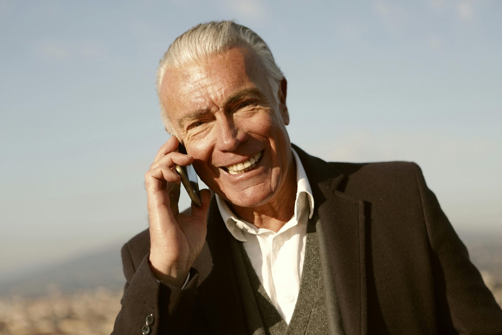 Senior man with grey hair smiling while talking on the phone outdoors with a blurred cityscape background.